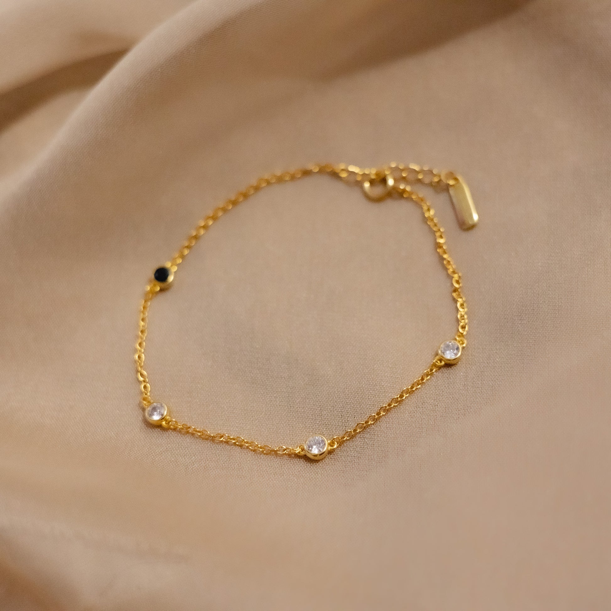 Delicate gold chain bracelet with clear bezel stones and one black accent stone on soft beige satin fabric.