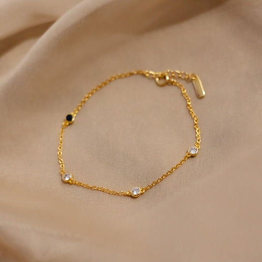 Delicate gold chain bracelet with clear bezel stones and one black accent stone on soft beige satin fabric.