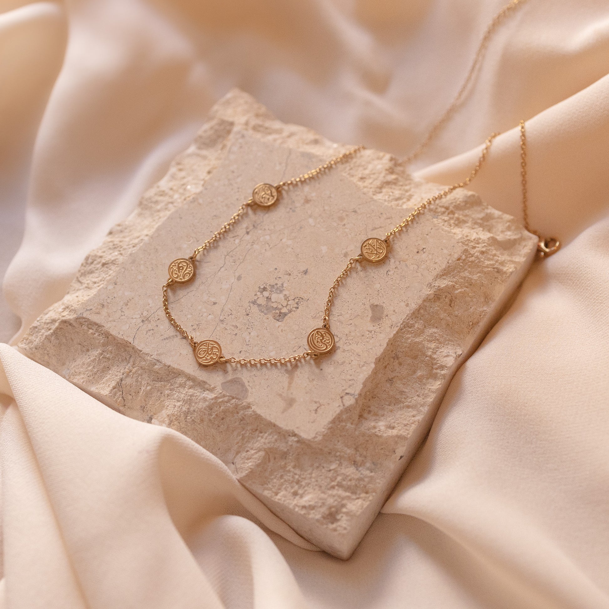 A gold chain bracelet inspired by the four corners of the Kaaba, featuring one black and three clear stones on a beige fabric background.