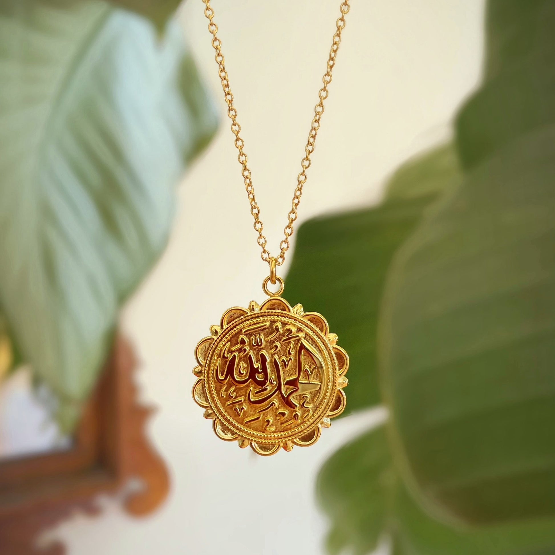 Alhamdulillah necklace gold with round Arabic calligraphy pendant and delicate chain displayed with green leaf background.