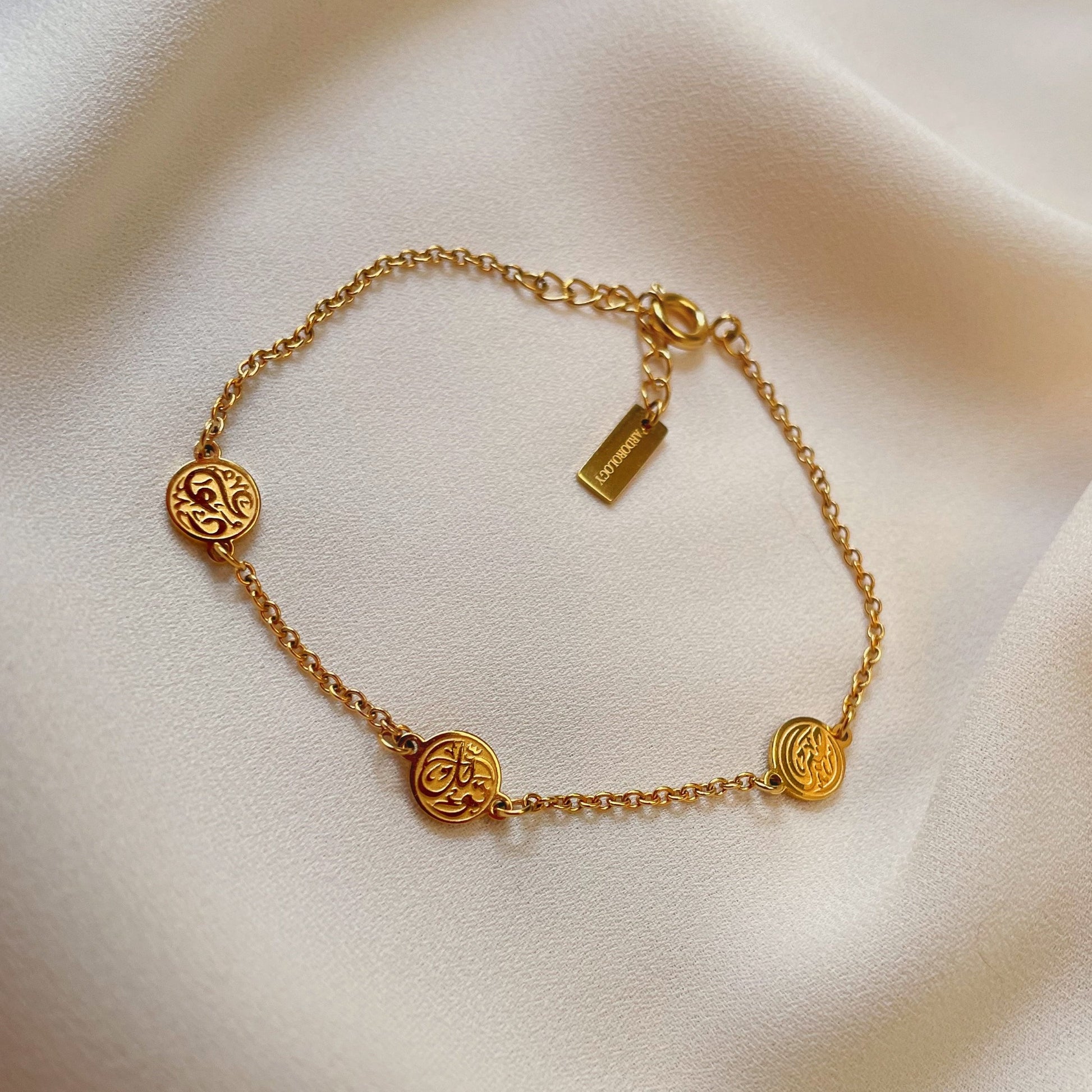 Gold hope coin bracelet with three round coin charms and delicate chain displayed on soft cream fabric.