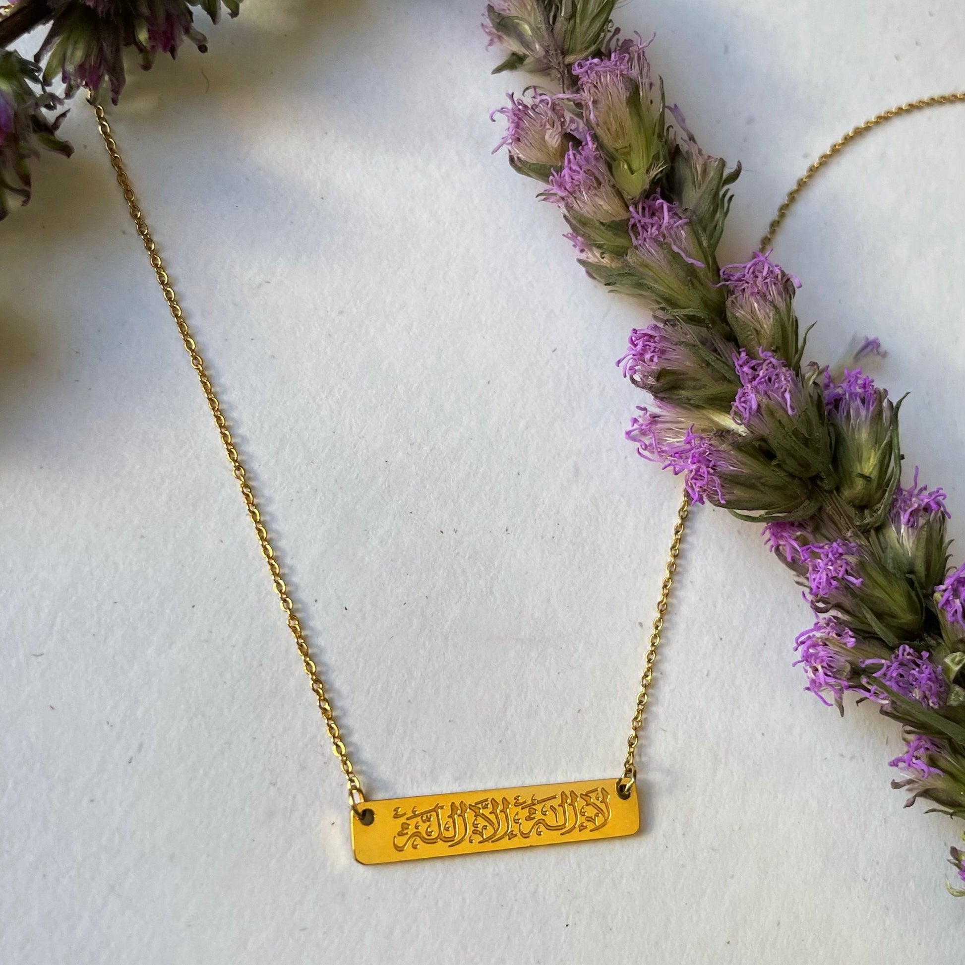 Gold La Ilaha Illallah necklace with Arabic calligraphy bar pendant and delicate chain displayed beside purple flowers.