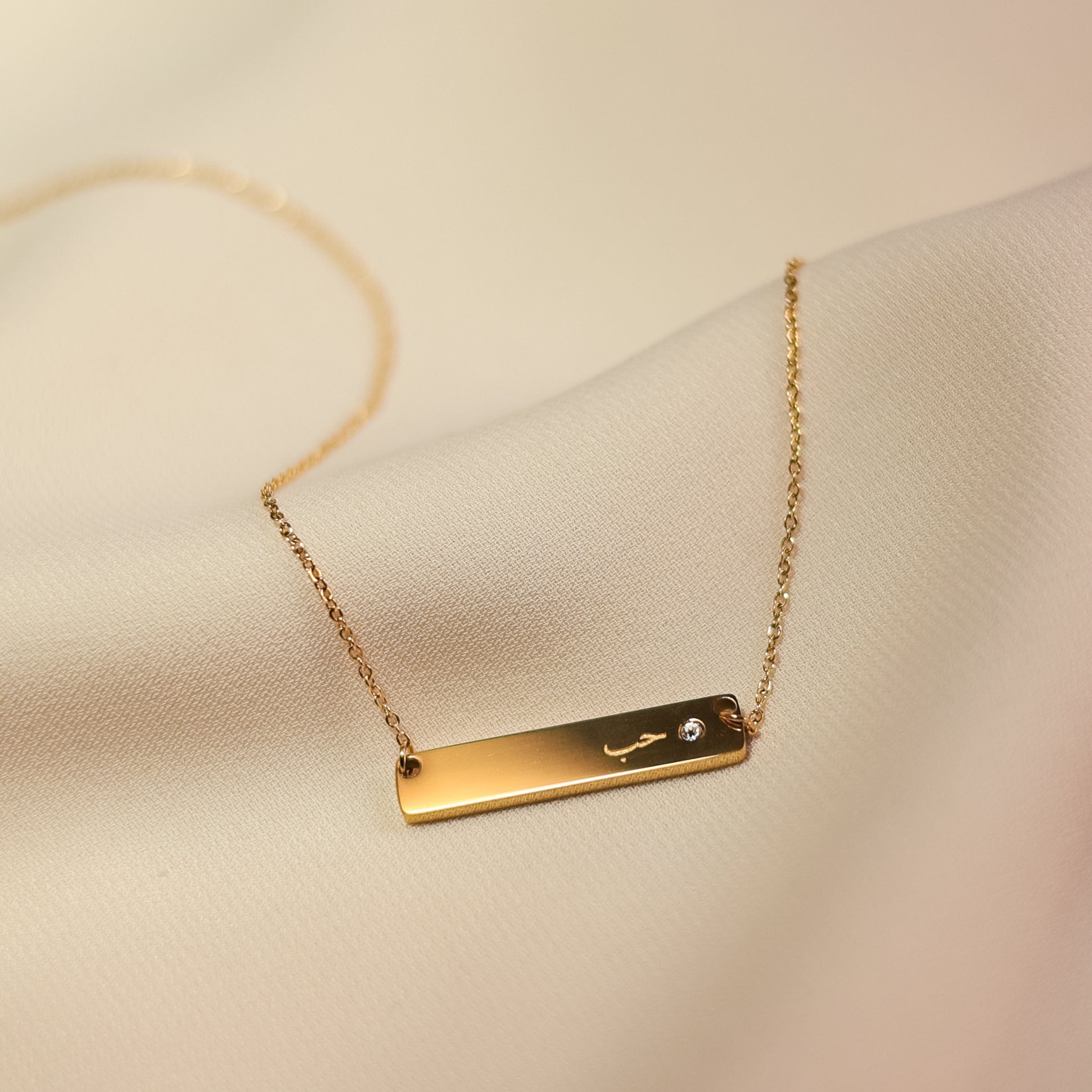 Love choker necklace gold with slim bar pendant, small crystal detail, and delicate chain on soft cream fabric.
