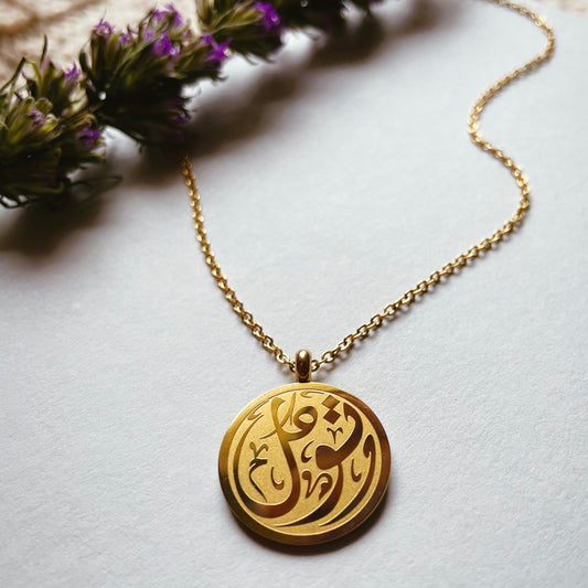 Tawakkul necklace gold with Arabic calligraphy pendant and delicate chain displayed on a soft cream background.